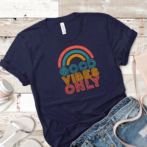 May include: Navy blue t-shirt with a retro rainbow design and the text "GOOD VIBES ONLY" in orange, teal, and yellow. The shirt is folded, with a pair of denim shorts, a pink purse, and white headphones in the background.