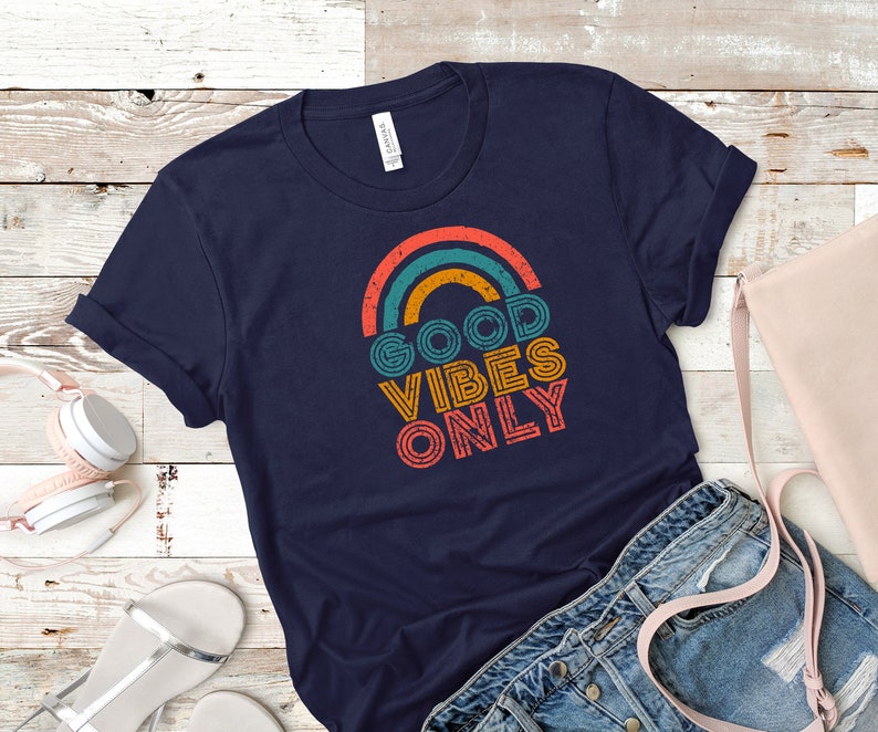 May include: Navy blue t-shirt with a retro rainbow design and the text "GOOD VIBES ONLY" in orange, teal, and yellow. The shirt is folded, with a pair of denim shorts, a pink purse, and white headphones in the background.