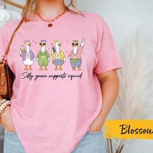 May include: A light pink short-sleeved t-shirt featuring a graphic of four cartoon geese wearing colorful outfits and sunglasses. The text below the geese reads "Silly goose supports squad". The geese are depicted with various accessories like a drink, a radio, and a fanny pack.