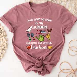 May include: A pink t-shirt with the text "I just want to work in my garden and hang out with my chickens". The shirt features a variety of gardening tools and chickens.