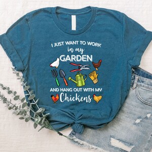 May include: A teal blue t-shirt with the text "I just want to work in my garden and hang out with my chickens". The shirt features a variety of gardening tools and chickens.