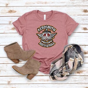 May include: A pink t-shirt with a black and white graphic of the band Aerosmith. The graphic features a winged heart with the band's name and the words "Authentic Rock & Roll".