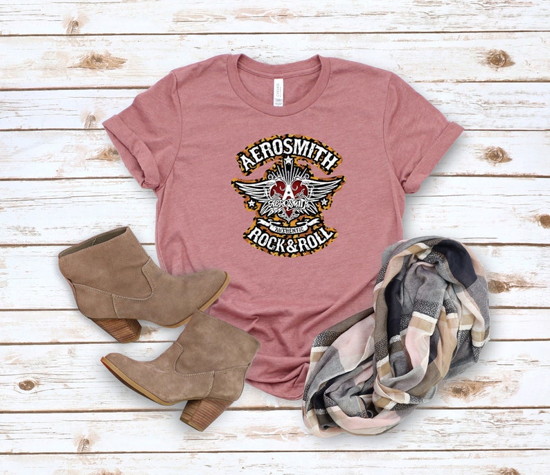 May include: A pink t-shirt with a black and white graphic of the band Aerosmith. The graphic features a winged heart with the band's name and the words "Authentic Rock & Roll".
