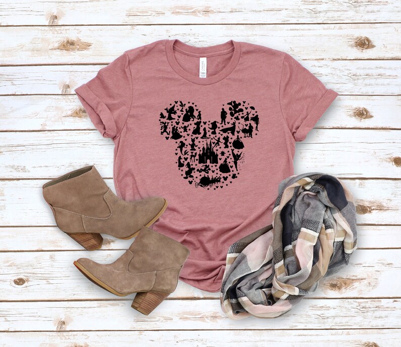 May include: Dusty rose t-shirt featuring a black silhouette of Mickey Mouse's head, composed of various Disney character icons. The shirt is paired with brown ankle boots and a plaid scarf. The shirt is made of a soft material.