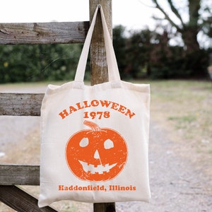 May include: A white canvas tote bag with an orange pumpkin design and the text "HALLOWEEN 1978 Haddonfield, Illinois".