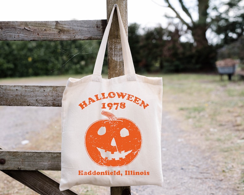May include: A white canvas tote bag with an orange pumpkin design and the text "HALLOWEEN 1978 Haddonfield, Illinois".