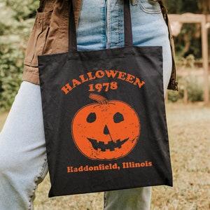 May include: A black tote bag with a white graphic of a pumpkin with a scary face. The text "HALLOWEEN 1978 Haddonfield, Illinois" is printed on the bag.