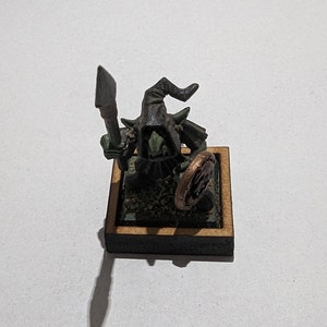 May include: A miniature green goblin figure wearing a dark hooded cloak, holding a grey spear in one hand and a round copper-colored shield in the other. The figure stands on a square base with a textured surface inside a wooden frame.