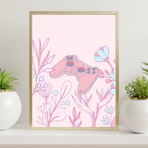 May include: A framed art print featuring a pink video game controller surrounded by pink and blue floral illustrations. The artwork has a gold frame and is set against a light pink background. Decorative plants in white pots flank the artwork.