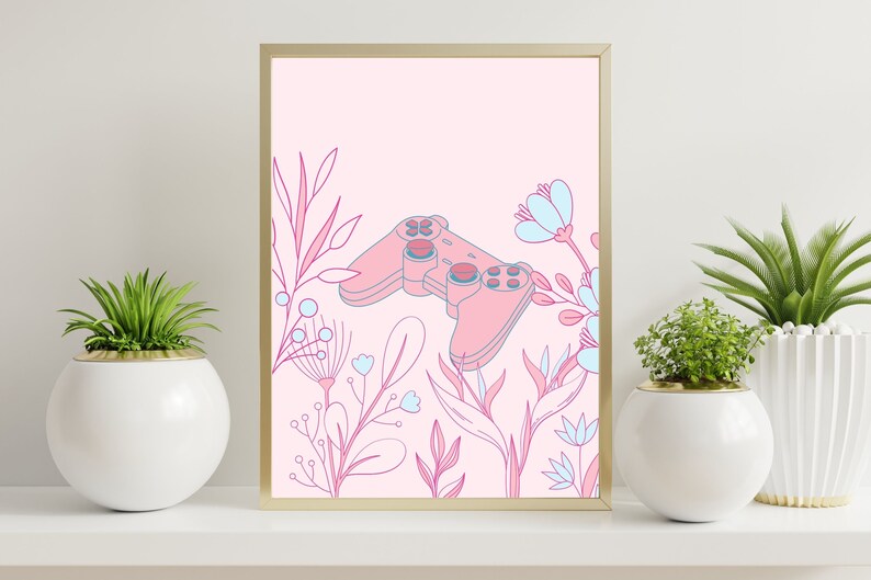 May include: A framed art print featuring a pink video game controller surrounded by pink and blue floral illustrations. The artwork has a gold frame and is set against a light pink background. Decorative plants in white pots flank the artwork.