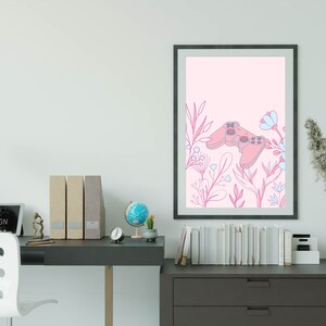 May include: A framed art print featuring a pink video game controller design with floral accents in pink and blue. The artwork is set against a light pink background. The print is displayed on a white wall above a desk.