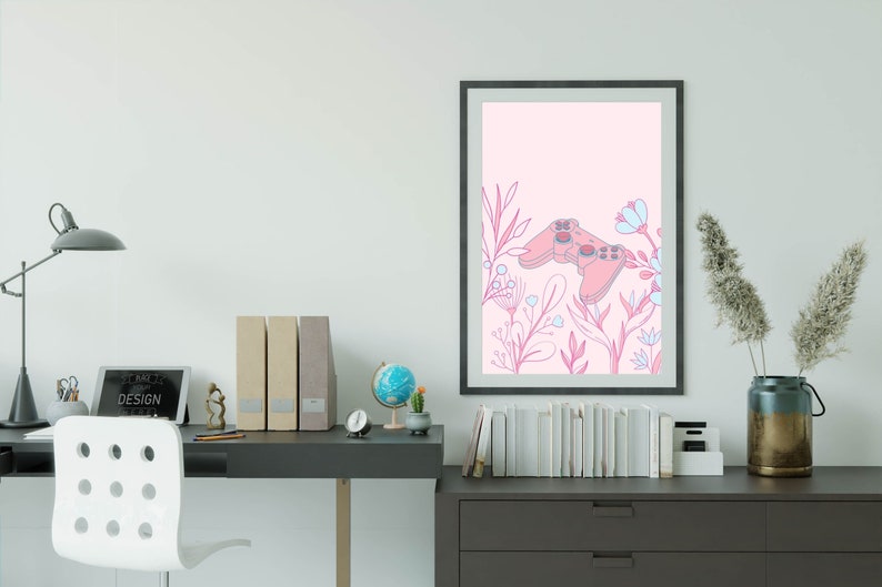 May include: A framed art print featuring a pink video game controller design with floral accents in pink and blue. The artwork is set against a light pink background. The print is displayed on a white wall above a desk.