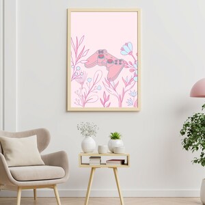 May include: A framed art print featuring a pink video game controller design surrounded by pink and blue floral elements. The artwork is set against a light pink background, creating a soft, feminine aesthetic. The frame is a light wood color.
