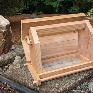 May include: A wooden bird feeder with a clear plastic front and a wire mesh bottom. The feeder has a hinged lid and a rope handle.