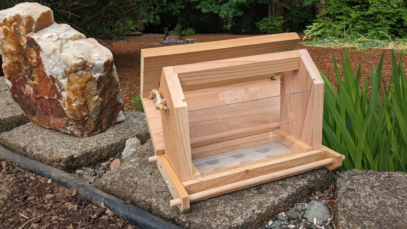 May include: A wooden bird feeder with a clear plastic front and a wire mesh bottom. The feeder has a hinged lid and a rope handle.