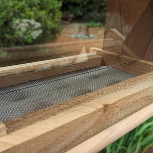 May include: A wooden bird feeder with a clear plastic top and a wire mesh bottom. The feeder has a wooden perch on the side.