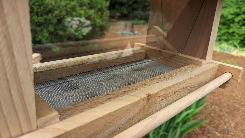 May include: A wooden bird feeder with a clear plastic top and a wire mesh bottom. The feeder has a wooden perch on the side.