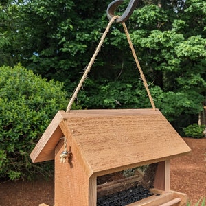 May include: A wooden bird feeder with a brown stain hanging from a metal hook. The feeder has a clear plastic window and a tray for seeds. The feeder is filled with black birdseed.