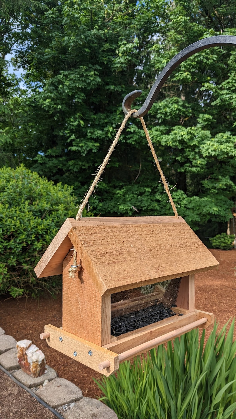 May include: A wooden bird feeder with a brown stain hanging from a metal hook. The feeder has a clear plastic window and a tray for seeds. The feeder is filled with black birdseed.