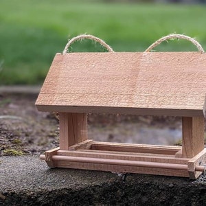 May include: A wooden bird feeder with a roof and a tray for birdseed. The feeder is made of natural wood and has a rustic look. It has two twine loops on the top for hanging.