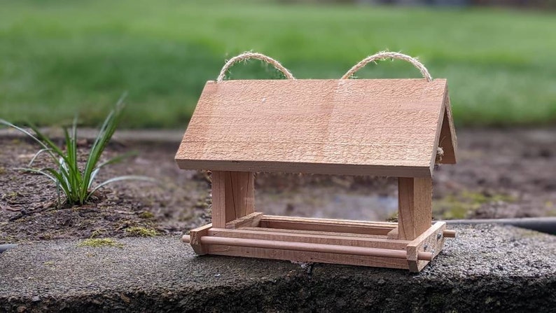 May include: A wooden bird feeder with a roof and a tray for birdseed. The feeder is made of natural wood and has a rustic look. It has two twine loops on the top for hanging.