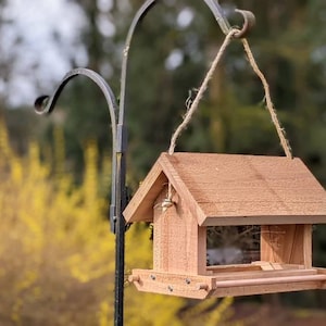 May include: A wooden bird feeder with a brown roof and a small perch. The feeder is hanging from a metal hook and is surrounded by a blurred background of yellow flowers and green trees.