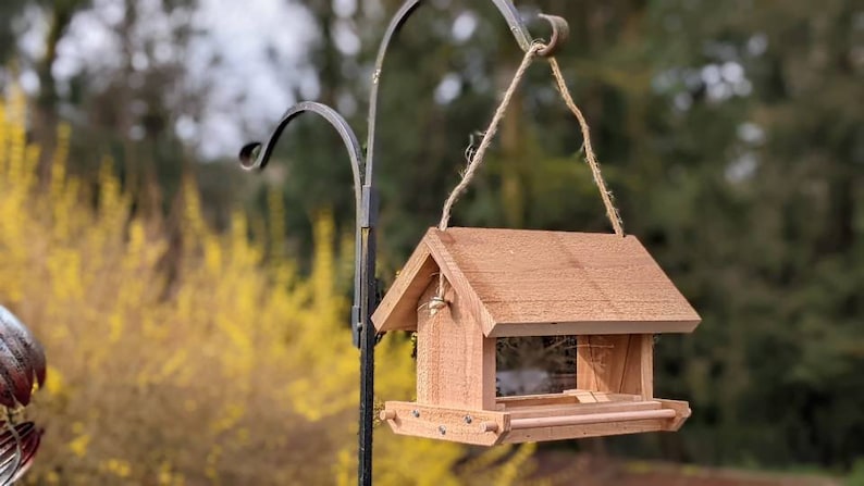 May include: A wooden bird feeder with a brown roof and a small perch. The feeder is hanging from a metal hook and is surrounded by a blurred background of yellow flowers and green trees.