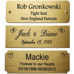 May include: Three gold-colored metal plaques with engraved text. The top plaque reads "Rob Gronkowski Tight End New England Patriots". The middle plaque reads "Jack & Diane September 13, 1988" with hearts on either side. The bottom plaque reads "Mackie Forever in our Hearts 03/16/1998-09/26/2012" with hearts on either side.