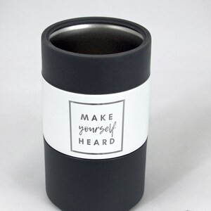 May include: A cylindrical tumbler with a black base and top, and a white band in the middle. The white band features the text "MAKE yourself HEARD" in a square frame. The tumbler has a silver interior.