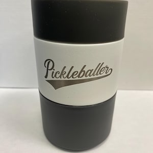 May include: A cylindrical tumbler with a black top and bottom, and a white center band. The word "Pickleballer" is printed in a script font on the white band. The tumbler is designed to hold beverages.