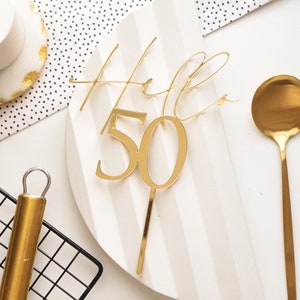 May include: Gold cake topper with the words "Hello 50" in a cursive font. The topper is on a white background.