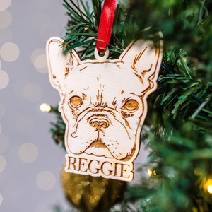 Op de afbeelding: Een houten kerstversiering met een Franse Bulldog met de naam "Reggie" gegraveerd op de onderkant. De versiering hangt aan een groene kerstboom met rood lint.