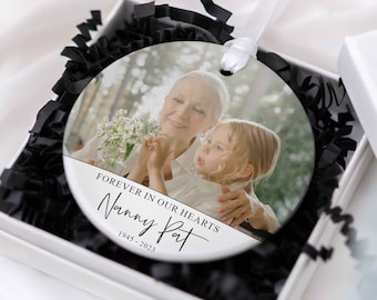 May include: A circular Christmas ornament featuring a photograph of a woman and child, displayed in a white gift box with black paper. The ornament has the text "Forever in our hearts, Nanny Pat, 1945-2023" and a white ribbon.