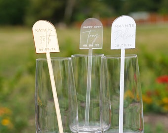 May include: Three champagne flutes with gold and white acrylic drink stirrers. The stirrers have personalized text, including "Katie's 30th 08.08.22", "Happy 50th Tony 08.08.22", and "Mr & Mrs Pulser 08.08.22".