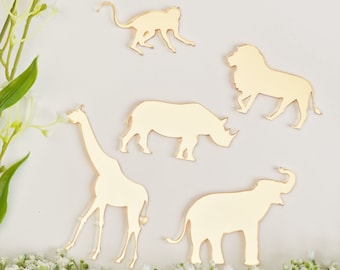 Può includere: Sagome di animali color oro tra cui una scimmia, un leone, un rinoceronte, una giraffa e un elefante. Le sagome sono disposte su una superficie bianca con accenti floreali, creando un tema safari o giungla.