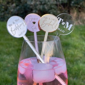 Könnte beinhalten: Drei runde Acryl-Getränkerührer mit personalisiertem Text. Die Rührer befinden sich in einem Glas mit rosa Getränk und Eis. Der Text auf den Rührern lautet "Mr & Mrs Bedding 24.07.22", "Emma & Sid" und "Mr & Mrs Birchall 20.6.22 T&M".