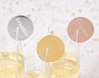 Op de afbeelding: Drie ronde, gespiegelde drankmarkeringen met de namen "Frankie", "Benjamin" en "Jason" erop gegraveerd. De markeringen zijn bevestigd aan heldere glazen drinkglazen met een zilveren, gouden en roségouden afwerking.