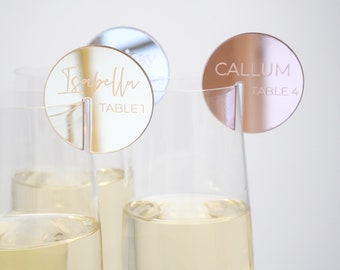 May include: Three champagne flutes with personalized acrylic table number place cards. The place cards are round and have a mirrored finish. The first place card says "Isabella Table 1" in gold lettering. The second place card says "Callum Table 4" in gold lettering. The third place card is silver.