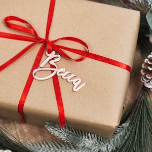 May include: A brown gift box wrapped with a red ribbon and a rose gold gift tag with the name "Becca" written on it.