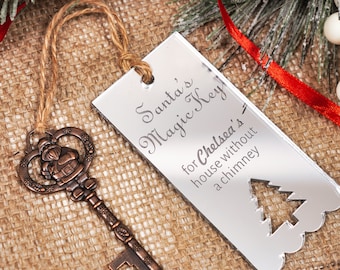 May include: A silver, rectangular ornament with the text "Santa's Magic Key for Chelsea's house without a chimney" and a cut-out Christmas tree. The ornament is hanging from a brown twine string. A vintage-style, bronze-colored key with a Santa figure on the top is also pictured.