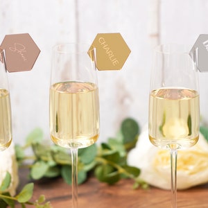 Op de afbeelding: Drie champagneflutes met gepersonaliseerde zeshoekige plaatskaarten. De kaarten zijn goud, rosé goud en zilver met de namen "Charlie", "Darren" en "Shane" erop.