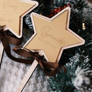 以下が含まれることがあります： 2つの星型のクリスマスオーナメントのクローズアップ。オーナメントは金色の木製フレームと茶色のリボンが付いています。1つは「Bonnie Believes」、もう1つは「Penny is officially on the Nice List」と書かれています。