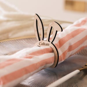 May include: A wooden bunny napkin ring with the name Callum engraved on it. The ring is holding a pink and white striped napkin.