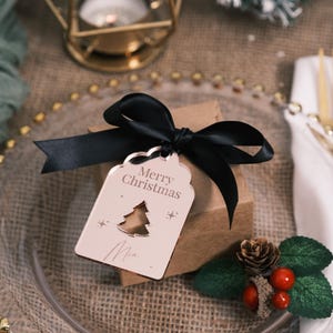 Puede incluir: Etiqueta de regalo de Navidad de oro rosa con un recorte de un árbol de Navidad y el texto "Merry Christmas" y "Mia". La etiqueta está atada con una cinta negra.