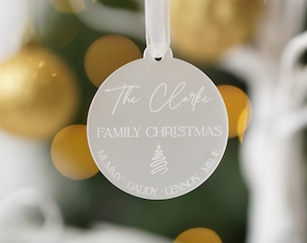 May include: A round, silver Christmas ornament with the text "The Clarke FAMILY CHRISTMAS" and names "Mummy, Daddy, Lennon, Milie". The ornament is hanging from a white ribbon, with a blurred background of gold Christmas lights.