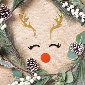May include: A Christmas wreath made of pine and eucalyptus branches with white berries. The wreath is decorated with gold reindeer antlers, black eyelashes, and a red nose.