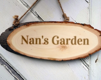 Könnte beinhalten: Ein rundes Holzschild mit rustikalem Aussehen hängt an einem Bindfaden. Das Schild ist aus einer Baumscheibe gefertigt und trägt die in goldenen Buchstaben gravierten Worte "Nan's Garden".