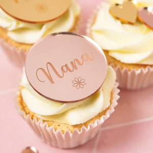 Könnte beinhalten: Ein Cupcake mit weißem Frosting und einer runden Scheibe in Roségold mit dem Wort "Nana" und einem kleinen Blumendesign.