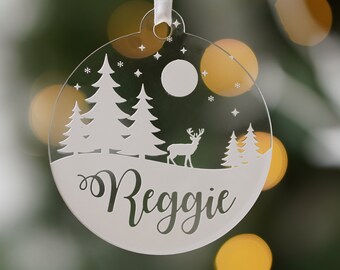 May include: A clear, circular Christmas ornament with a winter scene featuring trees, a deer, and the name "Reggie" in a script font. The ornament hangs from a white ribbon. Snowflakes and stars are also visible.
