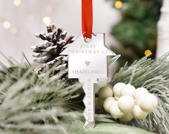 May include: Silver key-shaped Christmas bauble with a house design. The bauble reads "First Christmas at 1 Hazel Drive" with a heart symbol. The bauble is hanging from a red ribbon.